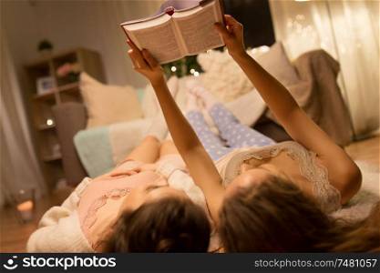 friendship, hygge and pajama party concept - happy female friends or teenage girls lying on floor and reading book at home. happy female friends reading book at home