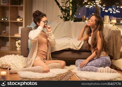 friendship, hygge and pajama party concept - happy young woman photographing her friend by vintage camera at home. female friends photographing by camera at home