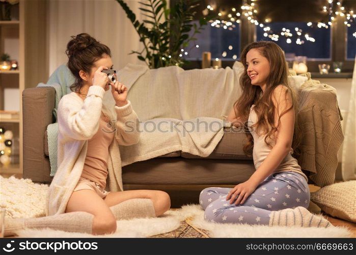 friendship, hygge and pajama party concept - happy young woman photographing her friend by vintage camera at home. female friends photographing by camera at home. female friends photographing by camera at home