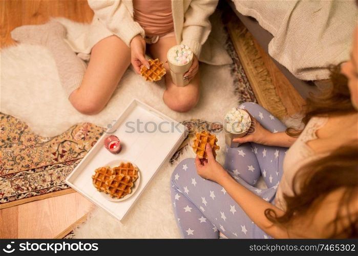 friendship, hygge and pajama party concept - two female friends or teenage girls drinking hot chocolate with marshmallow and eating waffles at home. women drinking hot chocolate and eating waffles