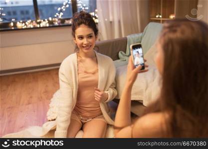 friendship, hygge and technology concept - happy young woman taking photo of her friend by smartphone at home. female friends photographing by smartphone at home. female friends photographing by smartphone at home