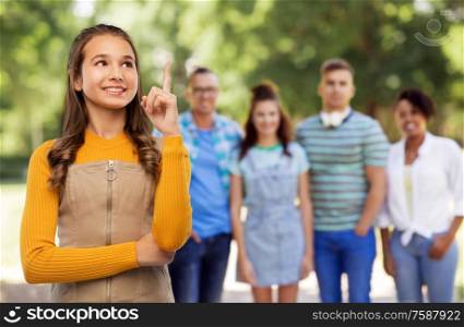 friendship, idea and people concept - happy smiling young teenage girl pointing finger up over group of friends at park on background. teenage girl pointing finger up over friends