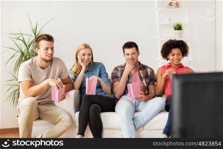friendship, junk food, people and entertainment concept - happy friends eating popcorn and watching tv at home