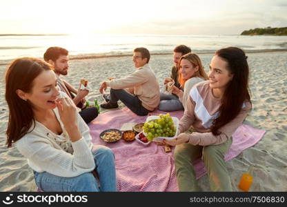 friendship, leisure and fast food concept - group of happy friends eating sandwiches or burgers at picnic on beach in summer. happy friends eating sandwiches at picnic on beach