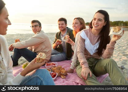 friendship, leisure and fast food concept - group of happy friends eating sandwiches or burgers at picnic on beach in summer. happy friends eating sandwiches at picnic on beach