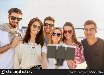 friendship, leisure and people concept - group of happy friends taking picture by smartphone on selfie stick on beach in summer. happy friends taking selfie on summer beach