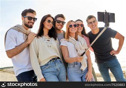 friendship, leisure and people concept - group of happy friends taking picture by smartphone on selfie stick on beach in summer. happy friends taking selfie on summer beach