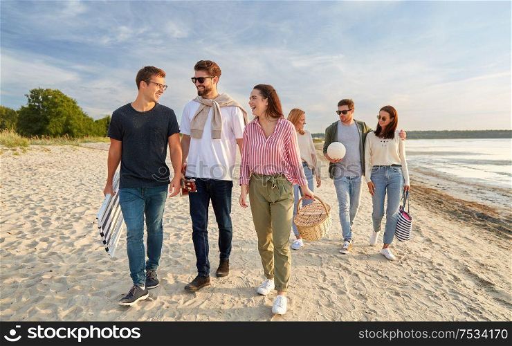 friendship, leisure and people concept - group of happy friends with ball, guitar, bag and picnic basket with blanket walking along beach in summer. happy friends walking along summer beach