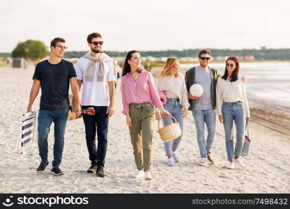 friendship, leisure and people concept - group of happy friends with guitar, chair and picnic basket walking along beach in summer. happy friends walking along summer beach