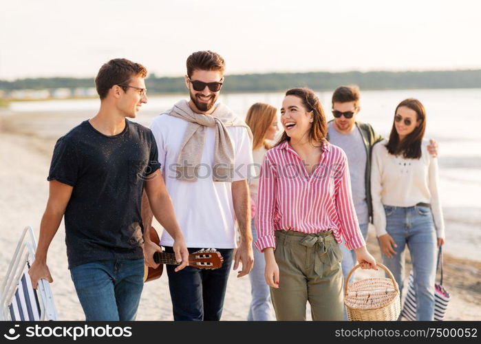 friendship, leisure and people concept - group of happy friends with guitar, chair and picnic basket walking along beach in summer. happy friends walking along summer beach