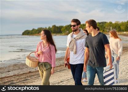 friendship, leisure and people concept - group of happy friends with guitar, picnic basket and folding chair walking along beach in summer. happy friends walking along summer beach