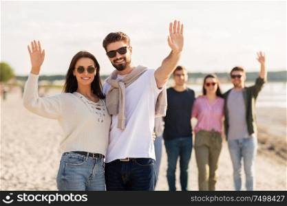 friendship, leisure and people concept - happy couple with group of friends on beach in summer waving hands. happy couple with friends waving hands on beach