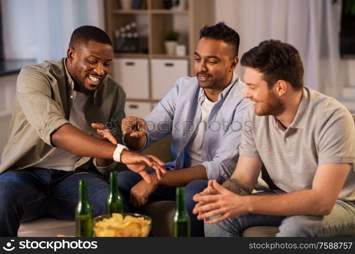 friendship, leisure and people concept - happy male friends with smart watch drinking beer and talking at home at night. male friends with smart watch and beer at home