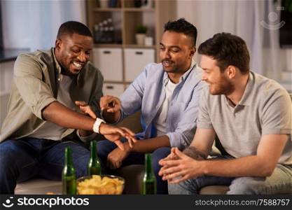 friendship, leisure and people concept - happy male friends with smart watch drinking beer and talking at home at night. male friends with smart watch and beer at home