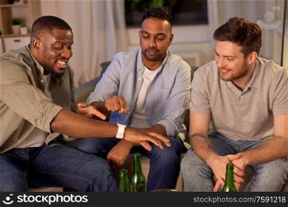 friendship, leisure and people concept - happy male friends with smart watch drinking beer and talking at home at night. male friends with smart watch and beer at home