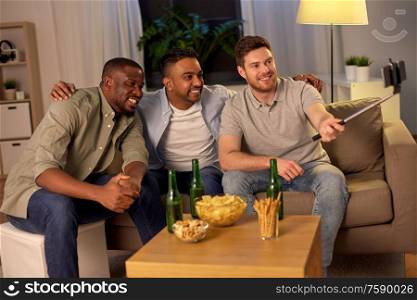 friendship, leisure and people concept - male friends taking picture with smartphone on selfie stick and drinking beer at home at night. male friends with smartphone taking selfie at home