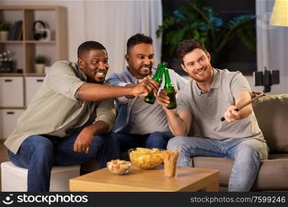 friendship, leisure and people concept - male friends taking picture with smartphone on selfie stick and drinking beer at home at night. male friends with smartphone taking selfie at home
