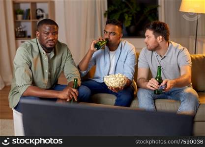 friendship, leisure and people concept - male friends with beer and popcorn watching tv at home at night. male friends with beer watching tv at home