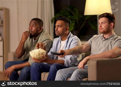 friendship, leisure and people concept - male friends with popcorn watching tv at home at night. male friends with popcorn watching tv at home