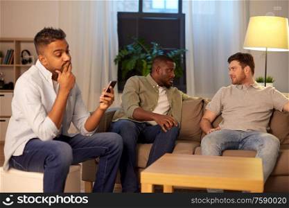 friendship, leisure and people concept - man using smartphone while his friends talking at home at night. man with smartphone and friends at home at night