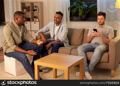 friendship, leisure and people concept - man using smartphone while his friends talking at home at night. man with smartphone and friends at home at night