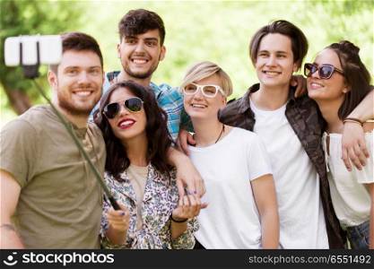 friendship, leisure and technology concept - group of happy smiling friends taking picture by selfie stick at summer park. friends taking picture by selfie stick at summer. friends taking picture by selfie stick at summer