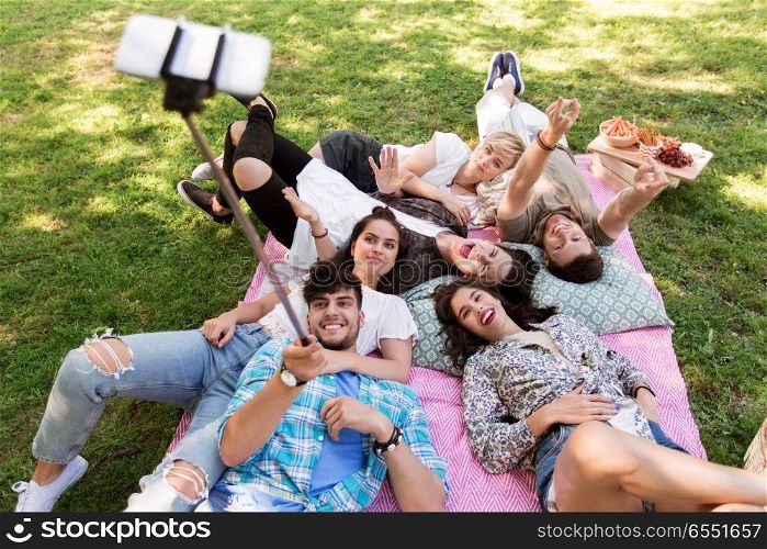 friendship, leisure and technology concept - group of happy smiling friends taking picture by selfie stick chilling on picnic blanket at summer park. friends taking picture by selfie stick at picnic. friends taking picture by selfie stick at picnic