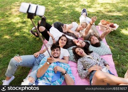 friendship, leisure and technology concept - group of happy smiling friends taking picture by selfie stick chilling on picnic blanket at summer park. friends taking picture by selfie stick at picnic. friends taking picture by selfie stick at picnic