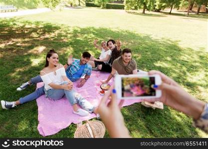 friendship, leisure and technology concept - group of happy smiling friends with non alcoholic drinks taking picture by smartphone on picnic at summer park. friends photographing by smartphone at picnic. friends photographing by smartphone at picnic