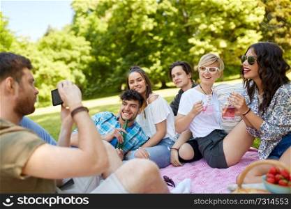 friendship, leisure and technology concept - guy taking picture on smartphone of friends drinking non alcoholic drinks at picnic in summer park. guy taking picture of friends on summer picnic