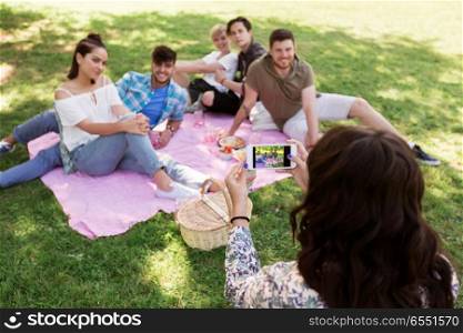 friendship, leisure and technology concept - woman taking picture of her friends by smartphone on picnic at summer park. woman picturing friends by smartphone at picnic. woman picturing friends by smartphone at picnic