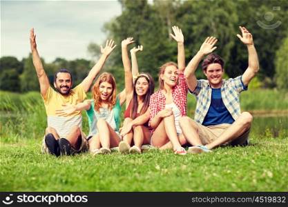 friendship, leisure, summer and people concept - group of smiling friends sitting on grass and waving hands outdoors