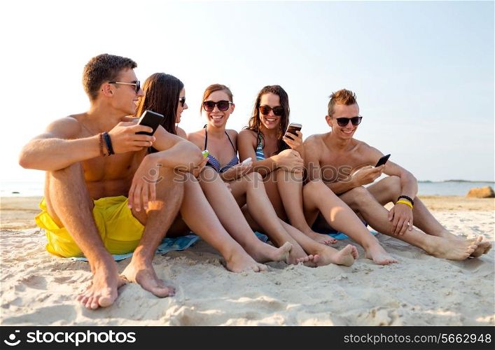 friendship, leisure, summer, technology and people concept - friends with smartphones sitting on sandy beach