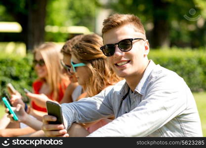 friendship, leisure, summer, technology and people concept - group of smiling friends with smartphones sitting on grass in park