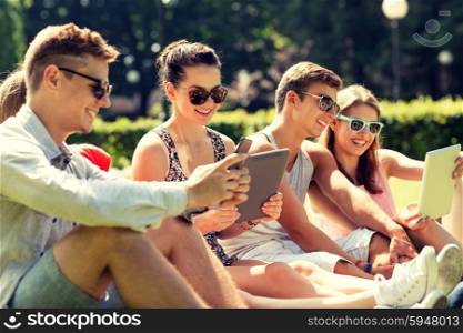 friendship, leisure, summer, technology and people concept - group of smiling friends with tablet pc computers and smartphone sitting on grass in park