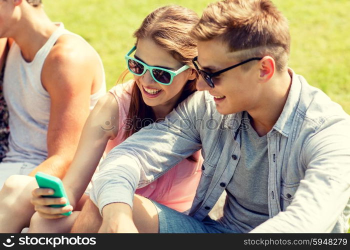 friendship, leisure, summer, technology and people concept - group of smiling friends with smartphone sitting on grass in park