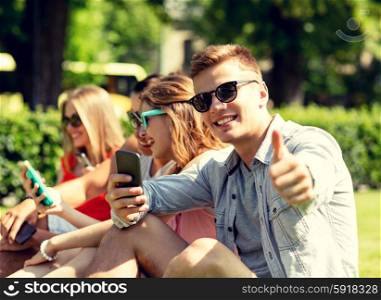 friendship, leisure, technology and people concept - smiling man with smartphones showing thumbs up in front of his friends