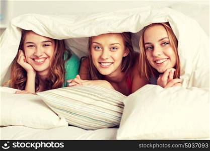 friendship, people and pajama party concept - happy friends or teenage girls having fun and lying under blanket with pillows in bed at home. happy young women in bed at home pajama party