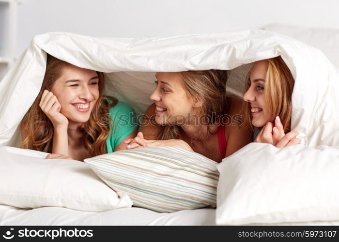 friendship, people and pajama party concept - happy friends or teenage girls having fun and lying under blanket with pillows in bed at home