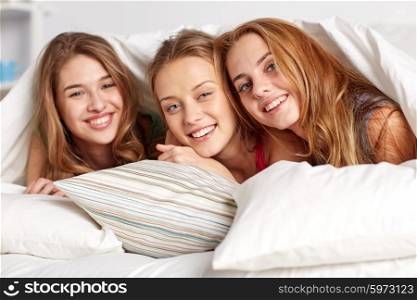 friendship, people and pajama party concept - happy friends or teenage girls having fun and lying under blanket with pillows in bed at home