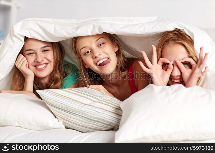 friendship, people and pajama party concept - happy friends or teenage girls having fun and lying under blanket with pillows in bed at home