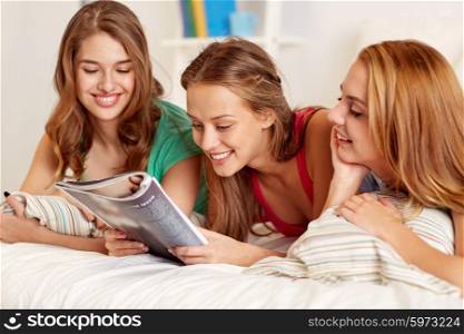 friendship, people and pajama party concept - happy friends or teenage girls reading magazine in bed at home