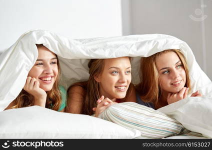 friendship, people and pajama party concept - happy friends or teenage girls having fun and lying under blanket with pillows in bed at home