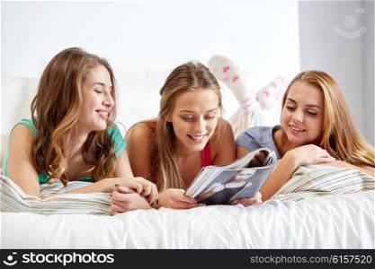 friendship, people and pajama party concept - happy friends or teenage girls reading magazine in bed at home