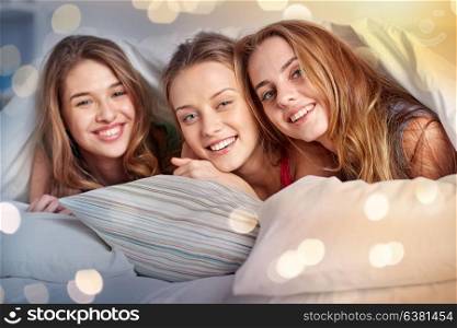 friendship, people and pajama party concept - happy friends or teenage girls having fun and lying under blanket with pillows in bed at home. happy young women in bed at home pajama party