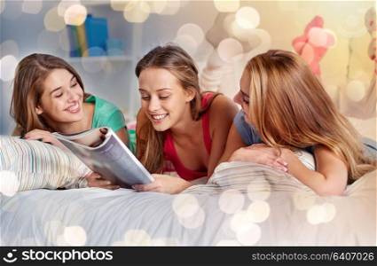 friendship, people and pajama party concept - happy friends or teenage girls reading magazine in bed at home. friends or teen girls reading magazine at home