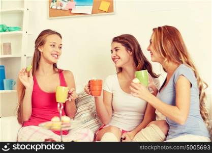 friendship, people, pajama party and communication concept - happy friends or teenage girls drinking tea and eating sweets at home. happy young women drinking tea with sweets at home
