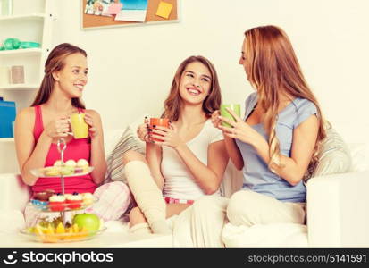 friendship, people, pajama party and communication concept - happy friends or teenage girls drinking tea and eating sweets at home. happy young women drinking tea with sweets at home