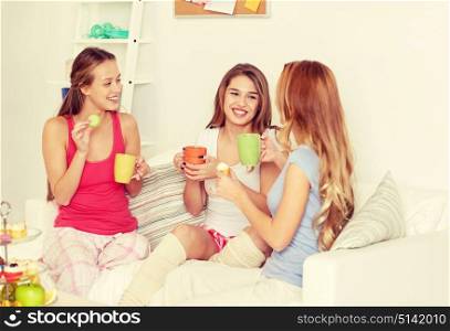 friendship, people, pajama party and communication concept - happy friends or teenage girls drinking tea and eating sweets at home. happy young women drinking tea with sweets at home