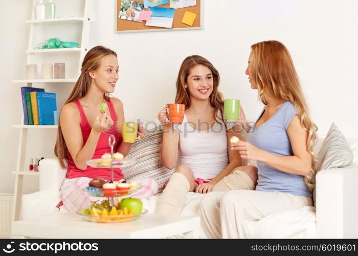 friendship, people, pajama party and communication concept - happy friends or teenage girls drinking tea and eating sweets at home